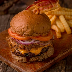 A pulled pork burger on a plank topped with onions and tomatoes and a pile of fries in the background.