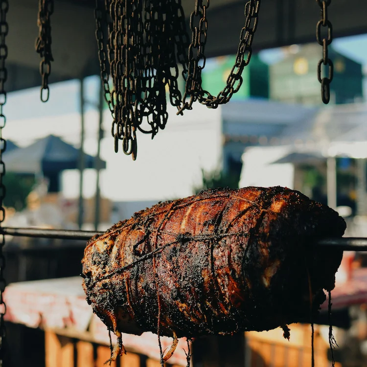 Charred pork shoulder wrapped with chord resting on a spit in front of a restaurant.