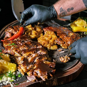 A pair of gloved hands organizing a platter of pulled pork and other assorted grilled sides.