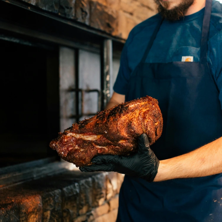 A man standing in front of a brick oven, wearing gloves and holding a spiced pork shoulder.