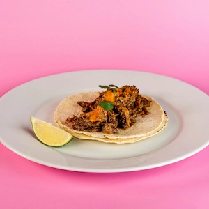 A pulled pork taco on a white plate with a lime wedge, in front of a pink background.